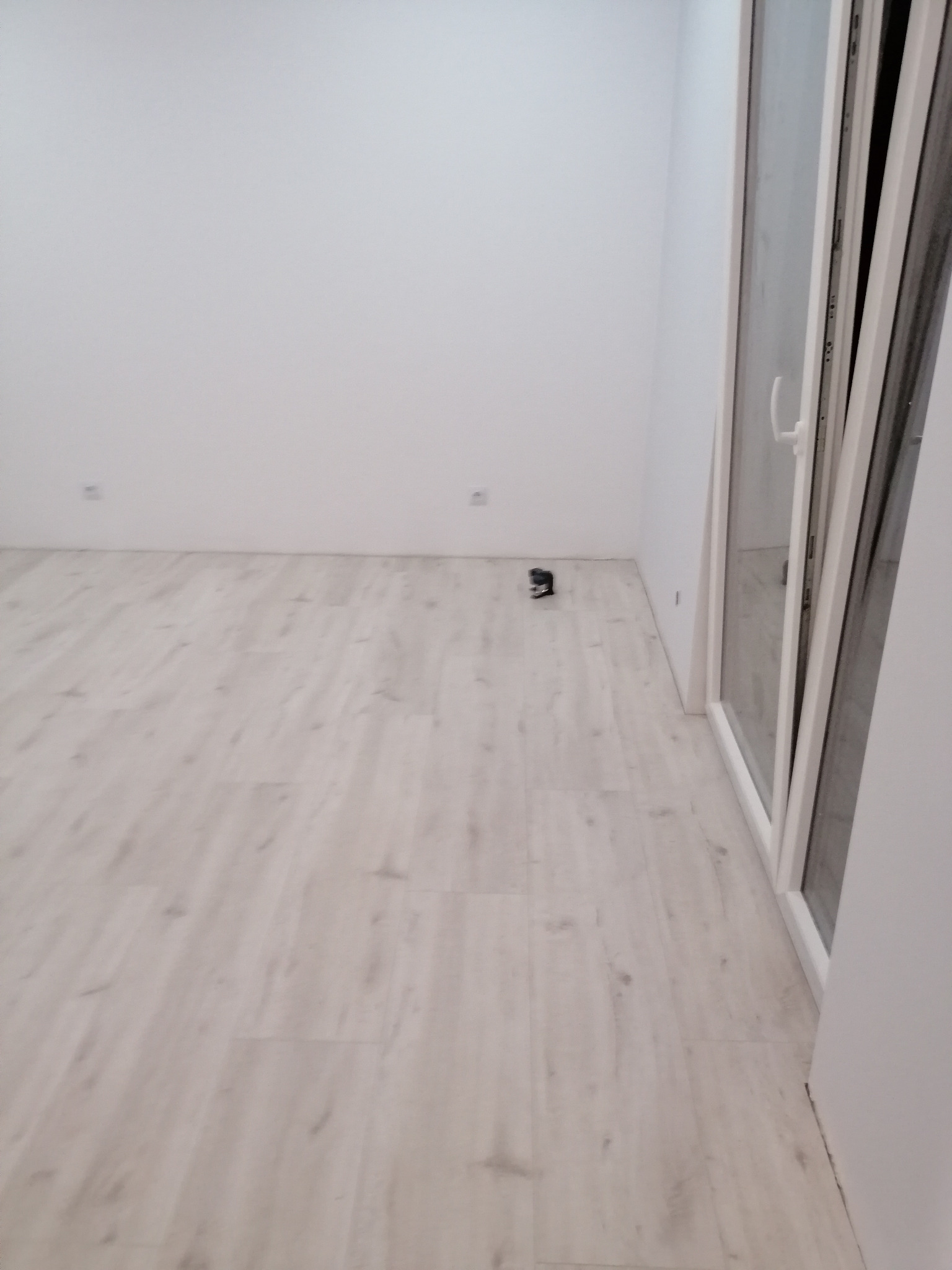 Jasne panele podłogowe ułożone w pustym, pomalowanym na biało pokoju, z widocznym poziomnicą laserową przy ścianie i oknem balkonowym.