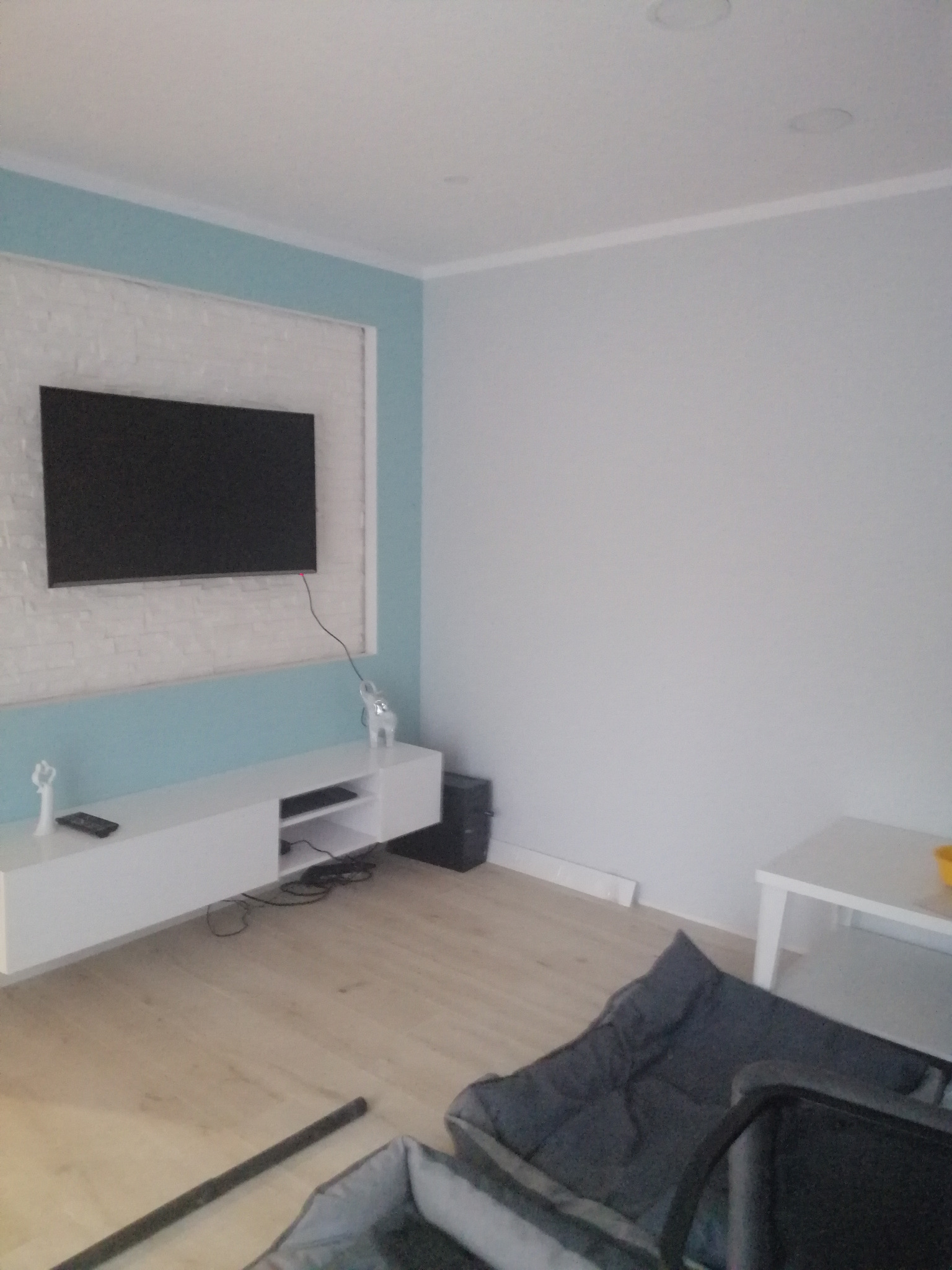 Telewizor na ścianie z białej cegły, podświetlonej turkusową farbą, biała szafka RTV z figurkami słonia i anioła, panele podłogowe, szara pufa i stolik w rogu pokoju.