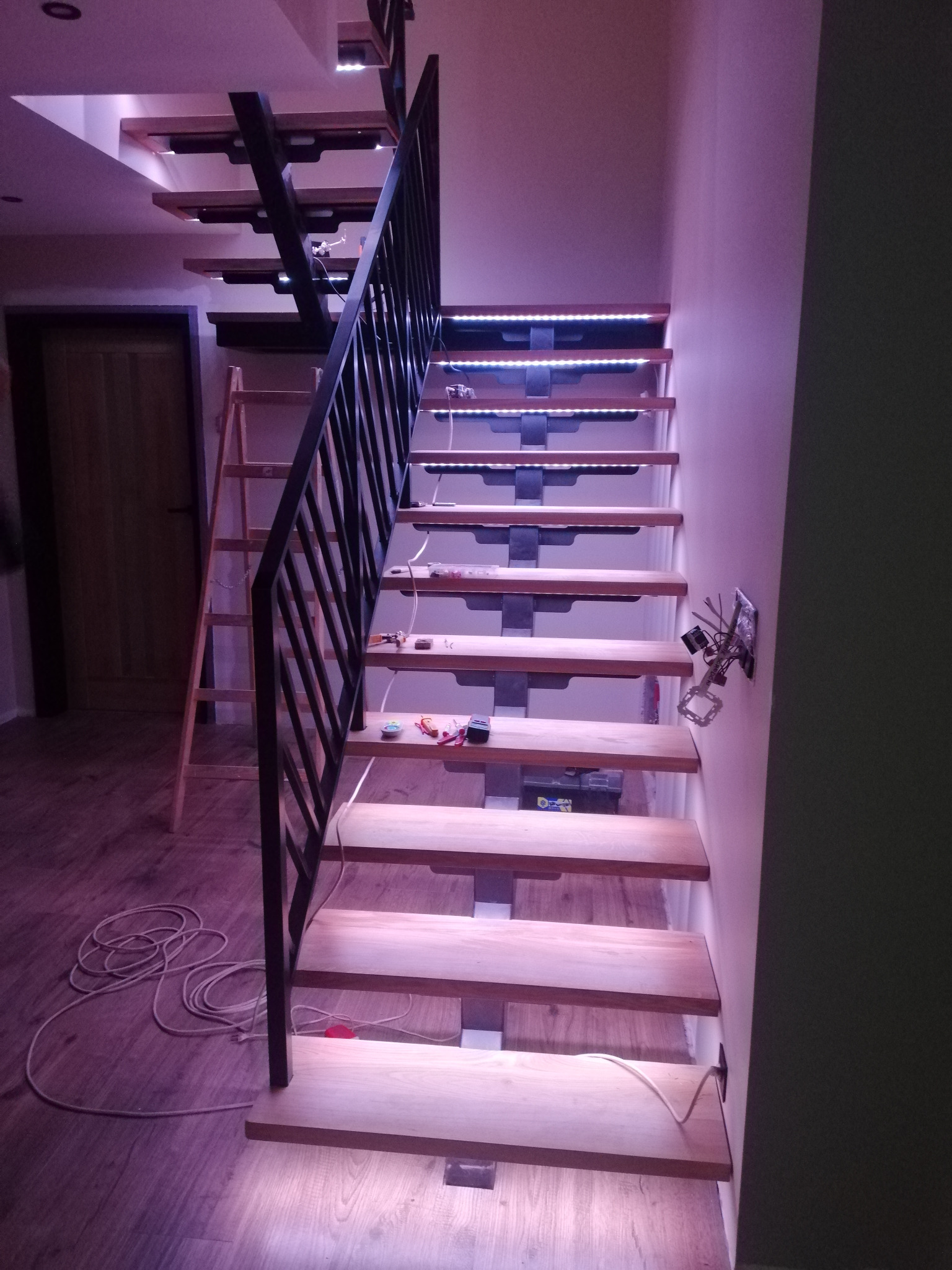 Schody drewniane z metalową barierką i podświetleniem LED w trakcie montażu, widoczne kable, skrzynka narzędziowa i drabina