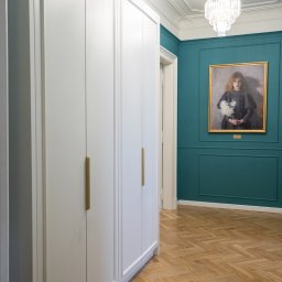 MCH Projekt Meble - Biała szafa na wymiar z długimi złotymi uchwytami, na tle turkusowej ściany z obrazem i parkietu w jodełkę, oświetlona kryształowymi żyrandolami.