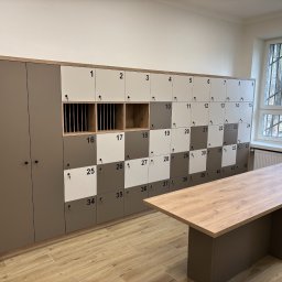 MCH Projekt Meble - Rząd szafek ubraniowych z numerami od 1 do 42, w kolorach szarym i białym, z drewnianymi wstawkami i stołem z drewnianym blatem na pierwszym planie, w jasnym pomieszczeniu z oknem z kratami.