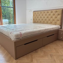 MCH Projekt Meble - Beżowe łóżko na wymiar z pikowanym zagłówkiem i szufladami, z szafką nocną w tym samym kolorze, na tle jasnego wnętrza z oknem i parkietem.