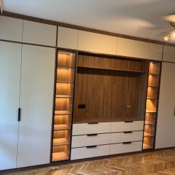 MCH Projekt Meble - Biało-drewniana szafa wnękowa z oświetleniem LED półek, widok z przodu na parkiet w jodełkę. Minimalistyczne uchwyty, ciepłe światło lampy sufitowej.