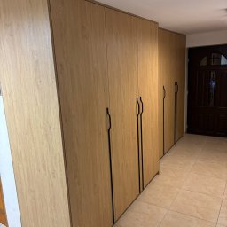MCH Projekt Meble - Długi rząd szaf na wymiar z drewnopodobnymi frontami i czarnymi, minimalistycznymi uchwytami, ustawionych wzdłuż korytarza z beżową podłogą.