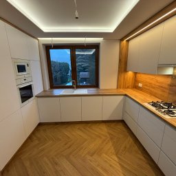 MCH Projekt Meble - Nowoczesna kuchnia z białymi frontami, drewnianym blatem i podłogą w jodełkę. Widoczne oświetlenie LED podszafkowe i okno z widokiem na zieleń.