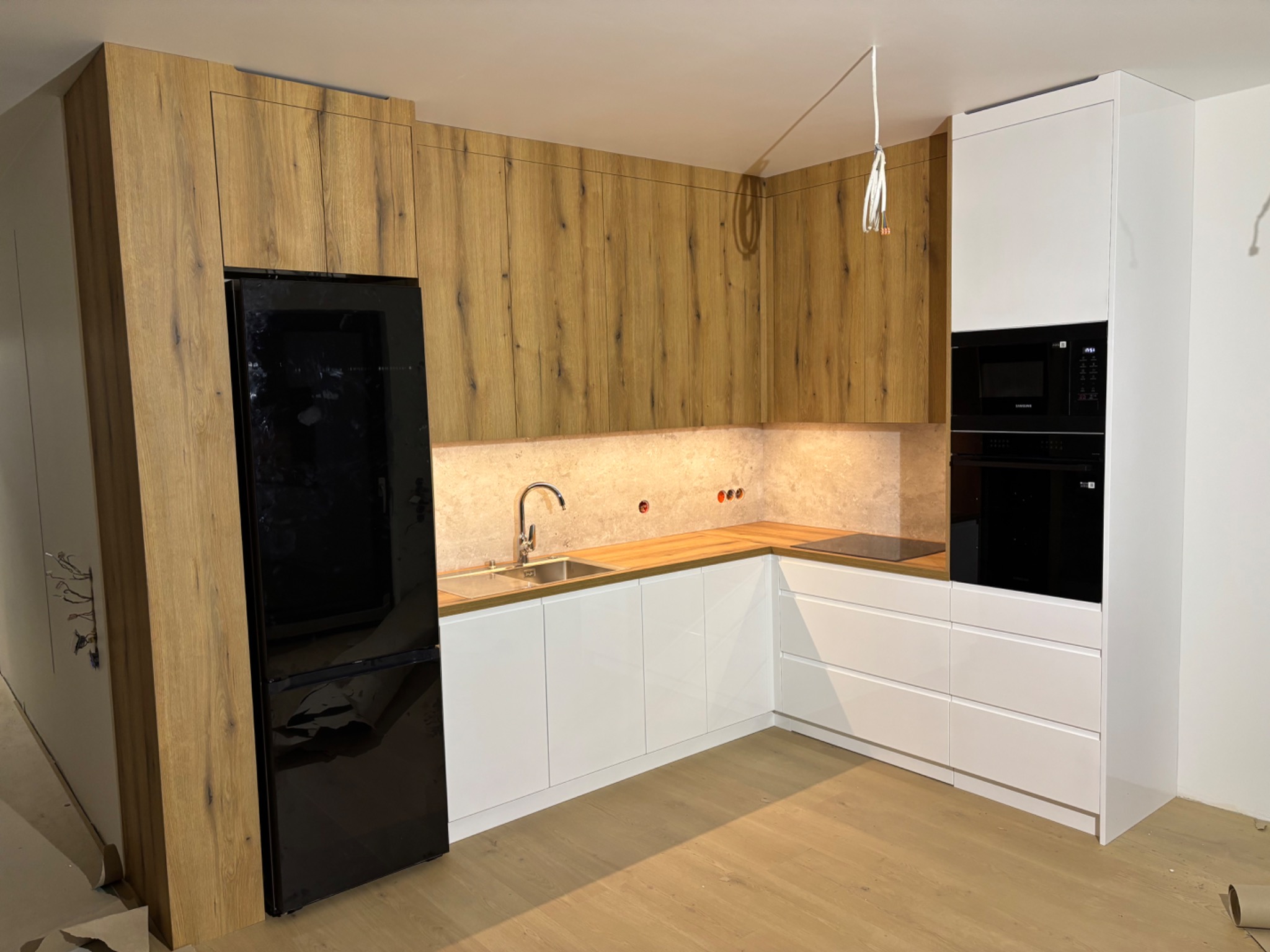 Nowoczesna kuchnia z drewnianymi szafkami górnymi i białymi dolnymi, czarna lodówka i sprzęt AGD w zabudowie, widoczne niedokończone prace instalacyjne.