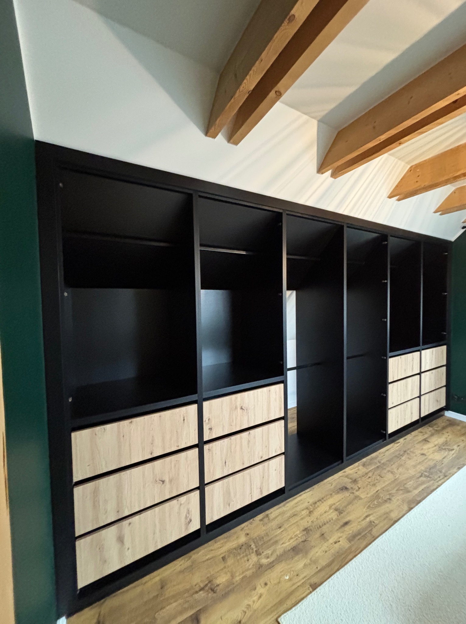 Czarna szafa wnękowa z drewnianymi szufladami, umieszczona w pomieszczeniu ze skośnym sufitem i drewnianymi belkami stropowymi, widoczna z boku.