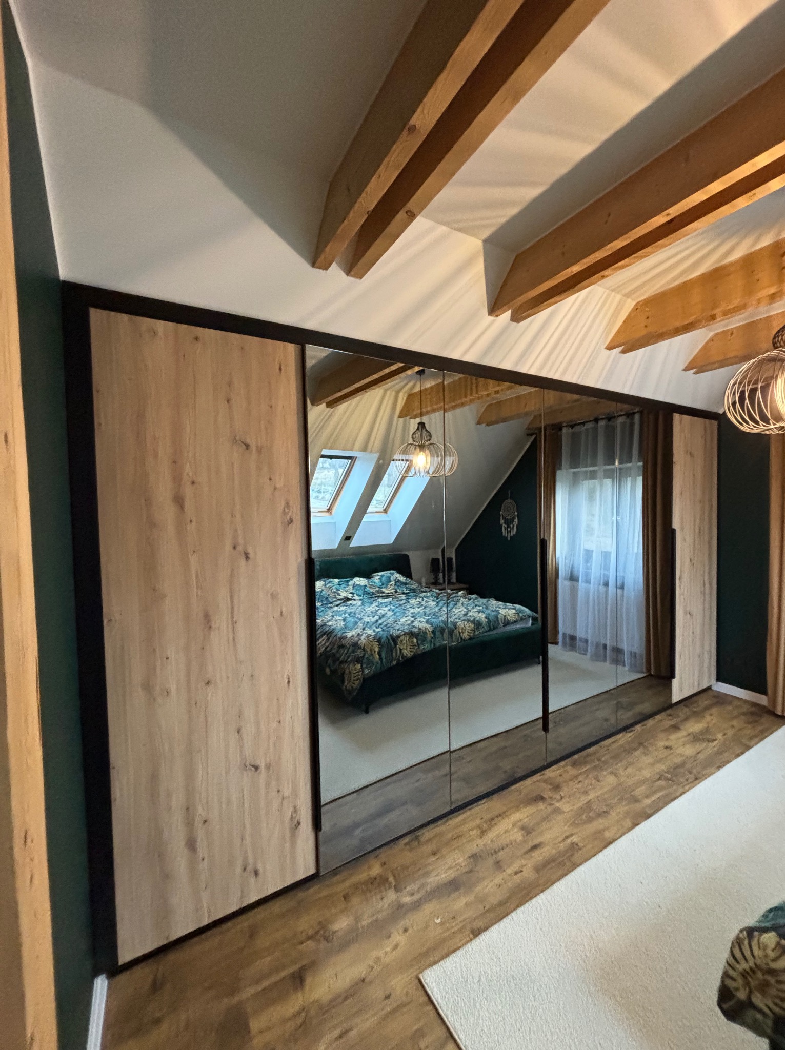 Szafa wnękowa z lustrami i drewnianymi panelami w sypialni na poddaszu z widoczną pościelą w kwiaty i drewnianymi belkami stropowymi.