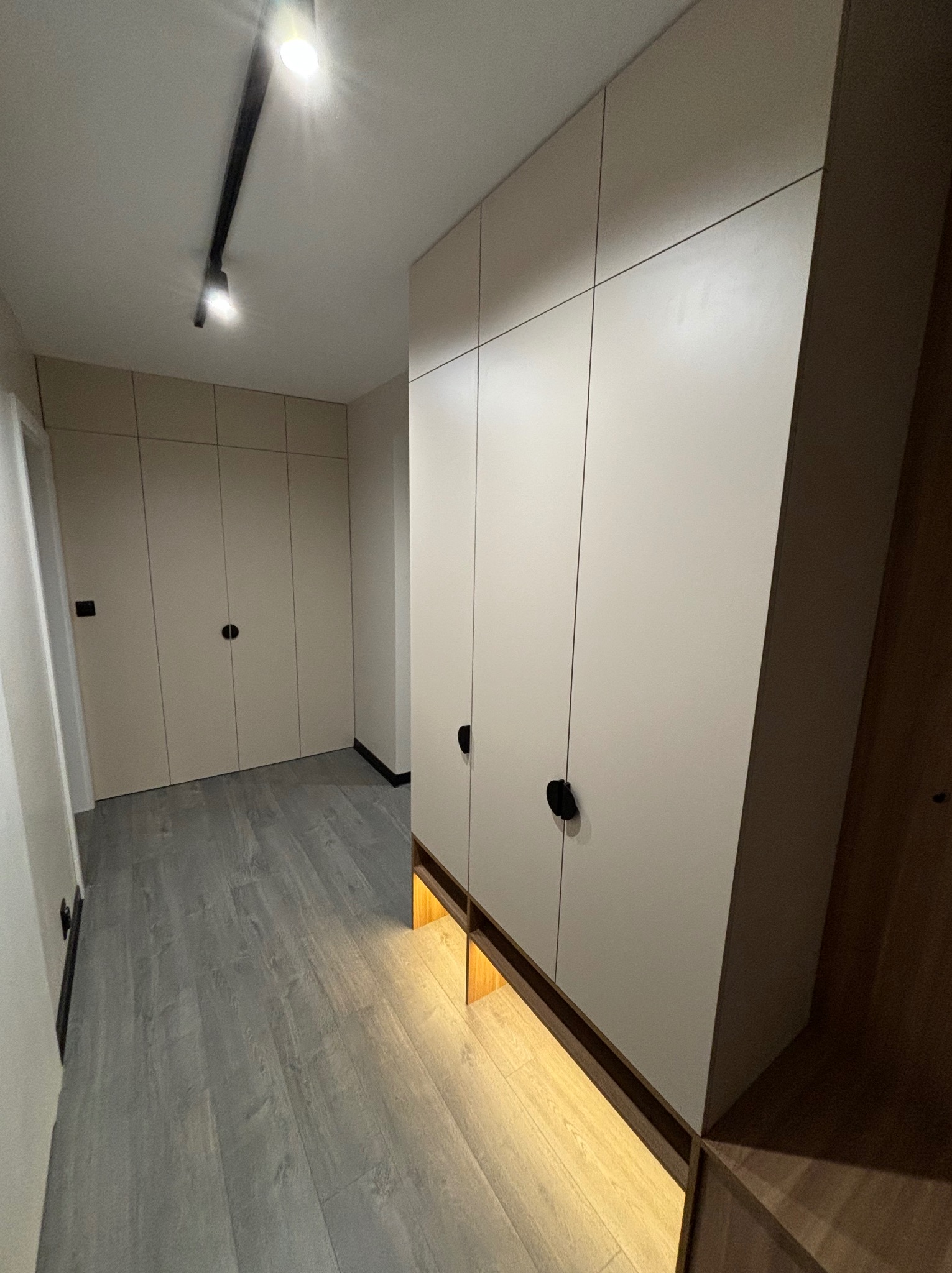 Biała szafa wnękowa z czarnymi, okrągłymi uchwytami, oświetlona od dołu ciepłym światłem, na szarej podłodze imitującej drewno.