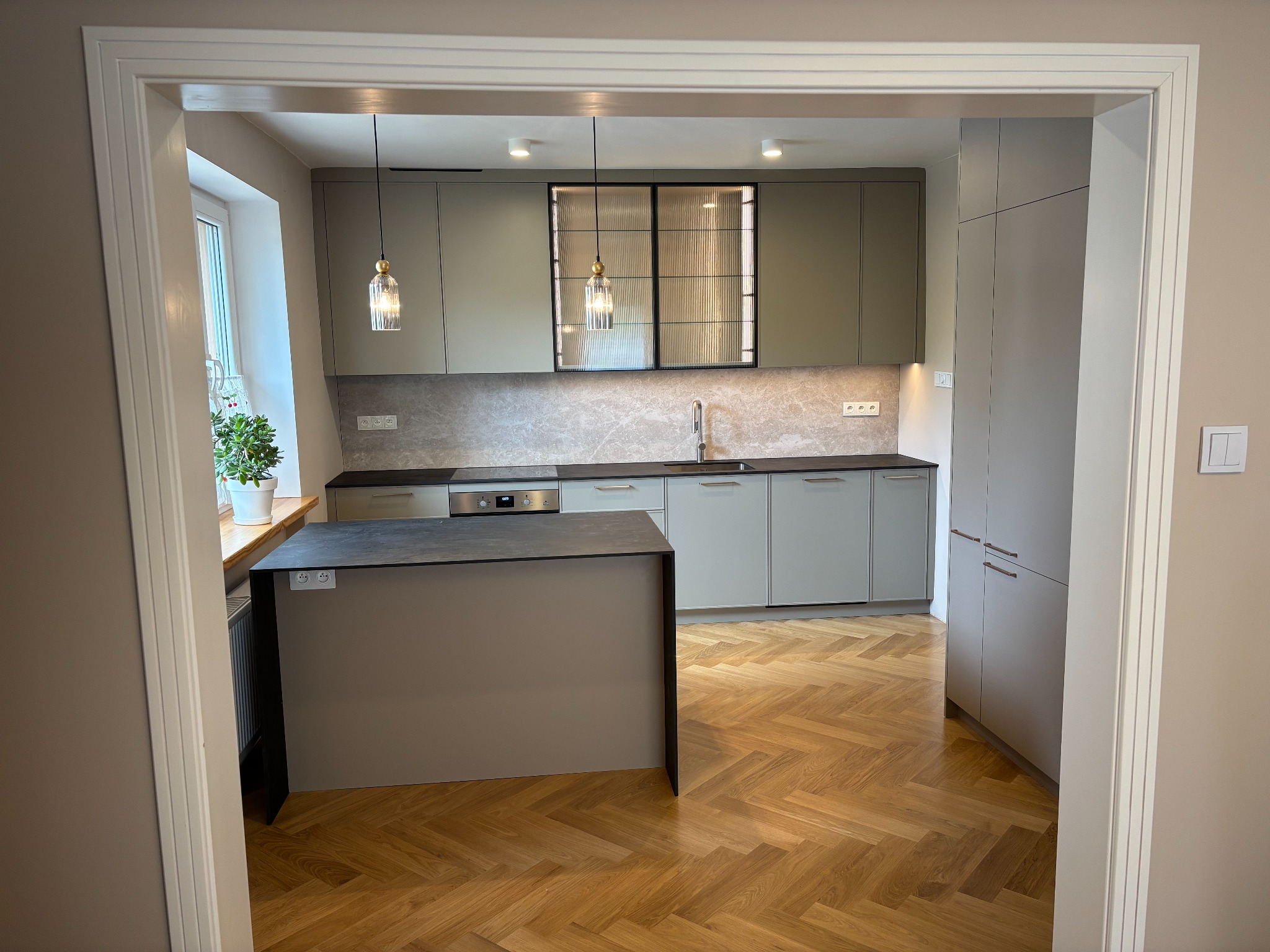 Jasnoszara kuchnia z wyspą, widziana z perspektywy przejścia. Parkiet w jodełkę, wiszące lampy i szafka z ryflowanym szkłem dodają elegancji.
