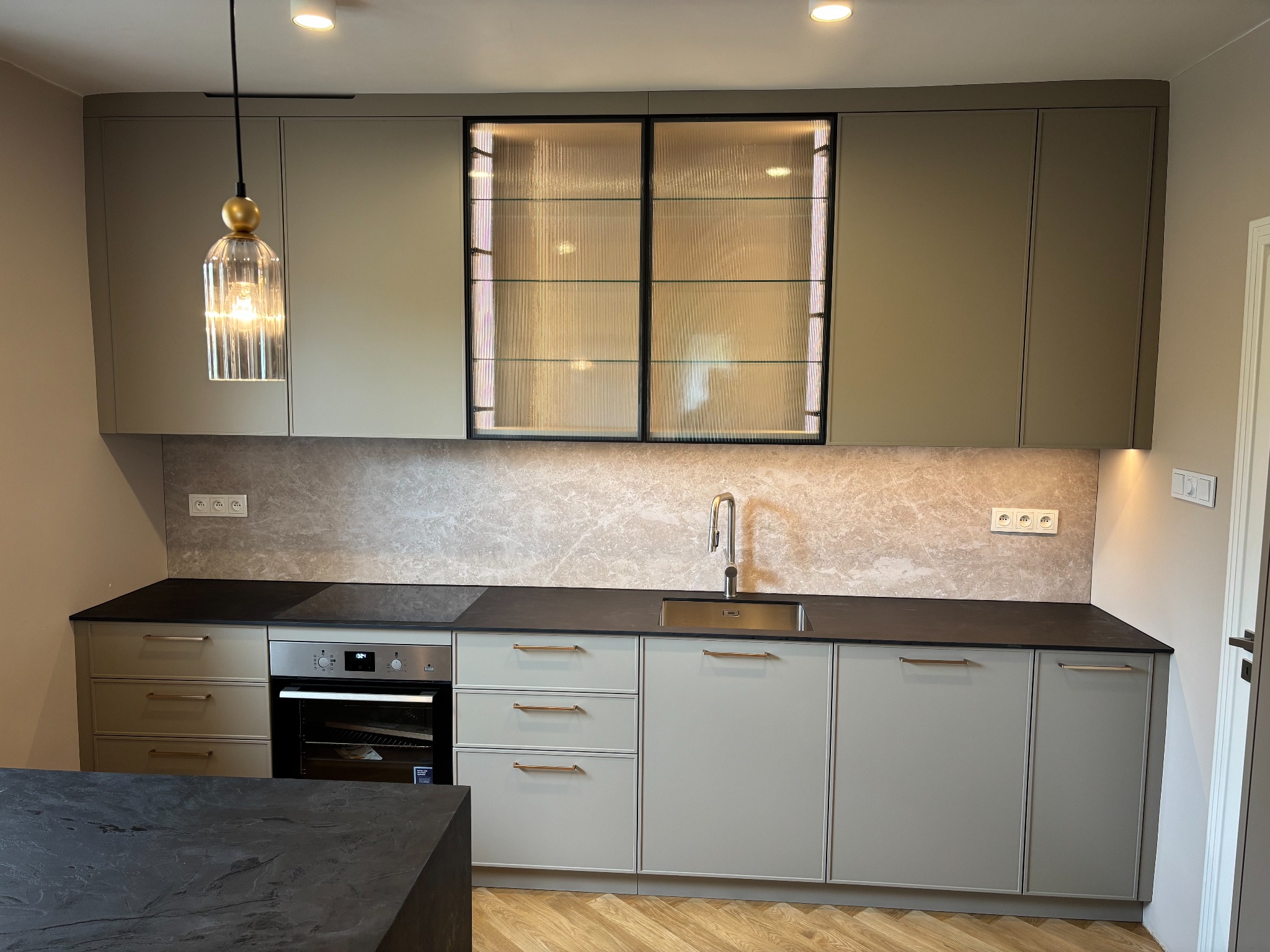 Nowoczesna kuchnia z szarymi frontami, czarnym blatem i marmurowym backsplashiem. Górna szafka z ryflowanym szkłem. Wisząca lampa z żarówką dekoracyjną.