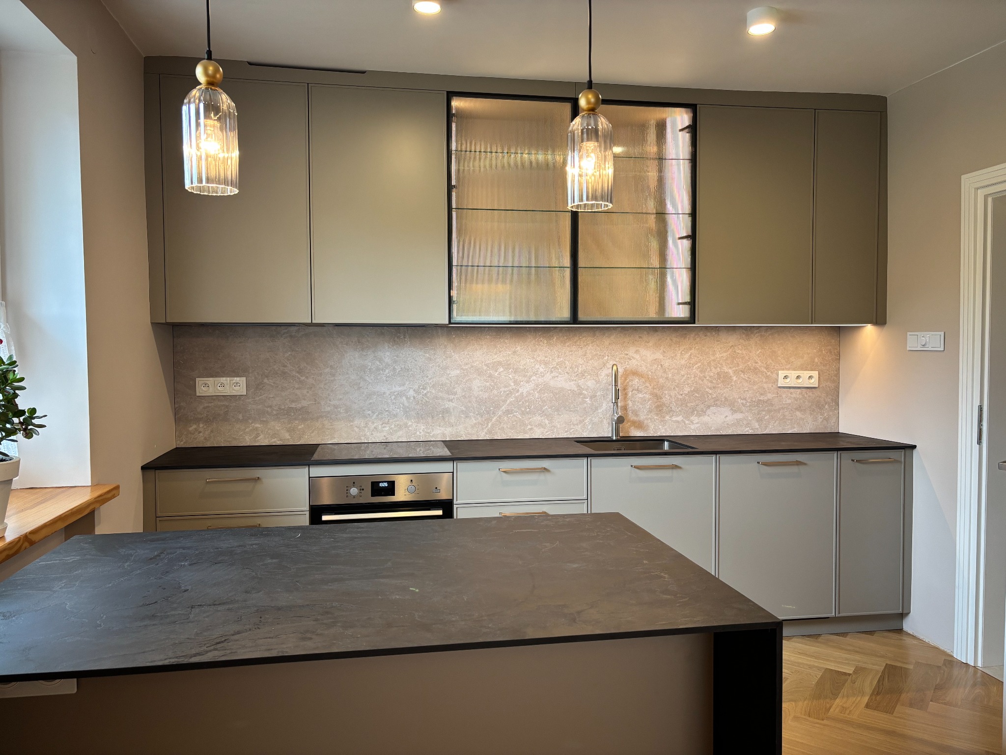 Nowoczesna kuchnia na wymiar w Krakowie z wyspą, fronty w odcieniach szarości, dekoracyjne lampy wiszące i szklane witryny w górnej zabudowie.