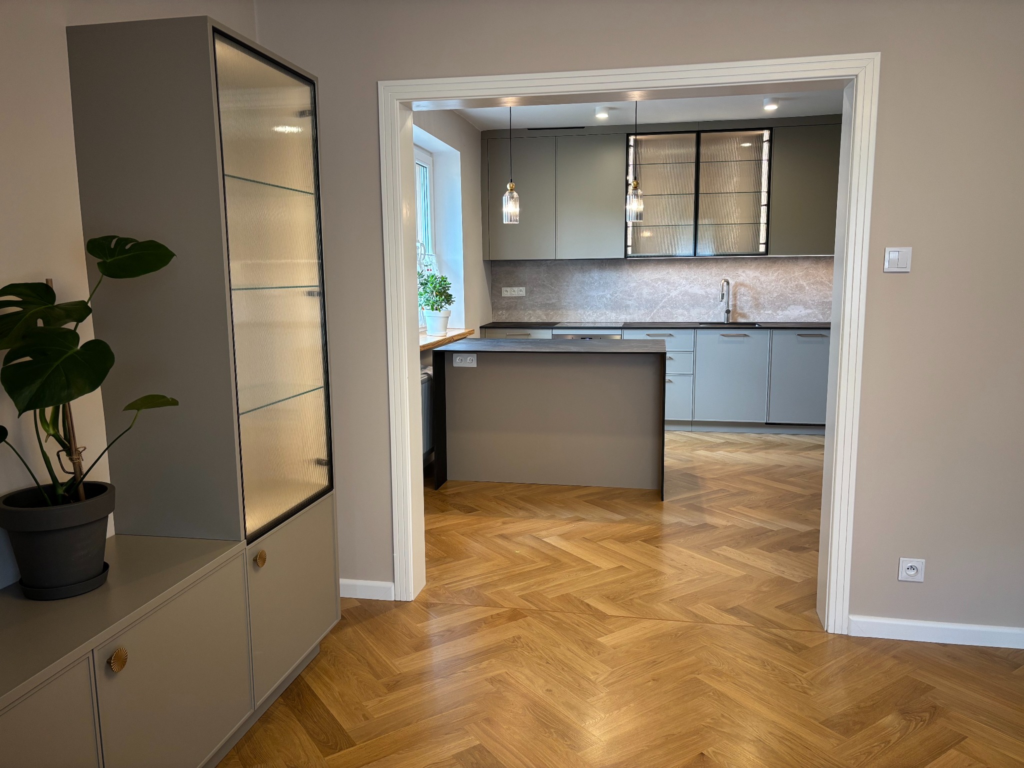 Nowoczesna kuchnia z wyspą, widziana z salonu. Parkiet w jodełkę, szare meble, oświetlenie punktowe i wiszące. Szafka z ryflowanymi szybami i rośliną.