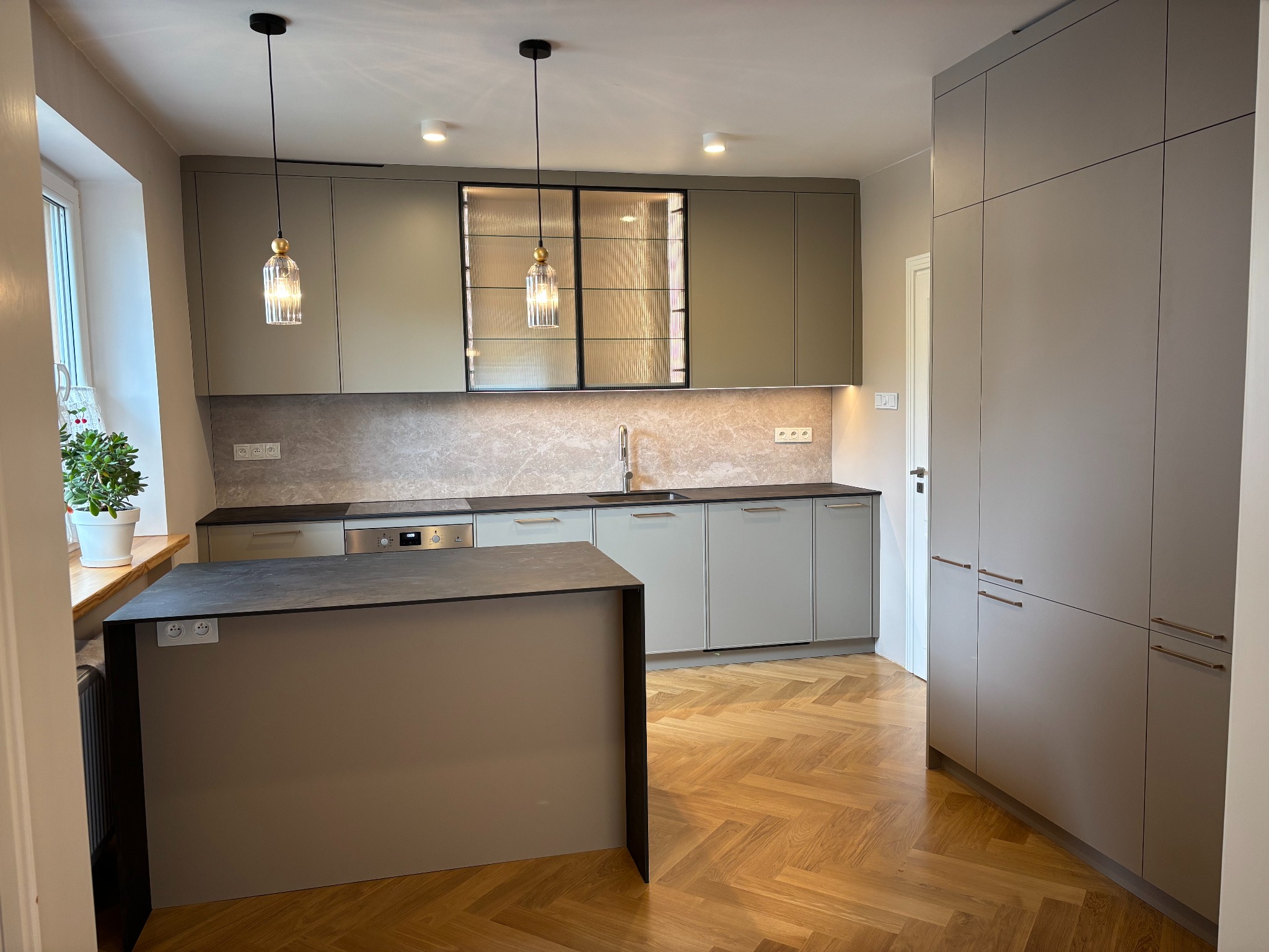 Nowoczesna kuchnia w odcieniach szarości z wyspą i parkietem w jodełkę. Uwagę przyciągają szklane fronty szafek wiszących i designerskie lampy wiszące.