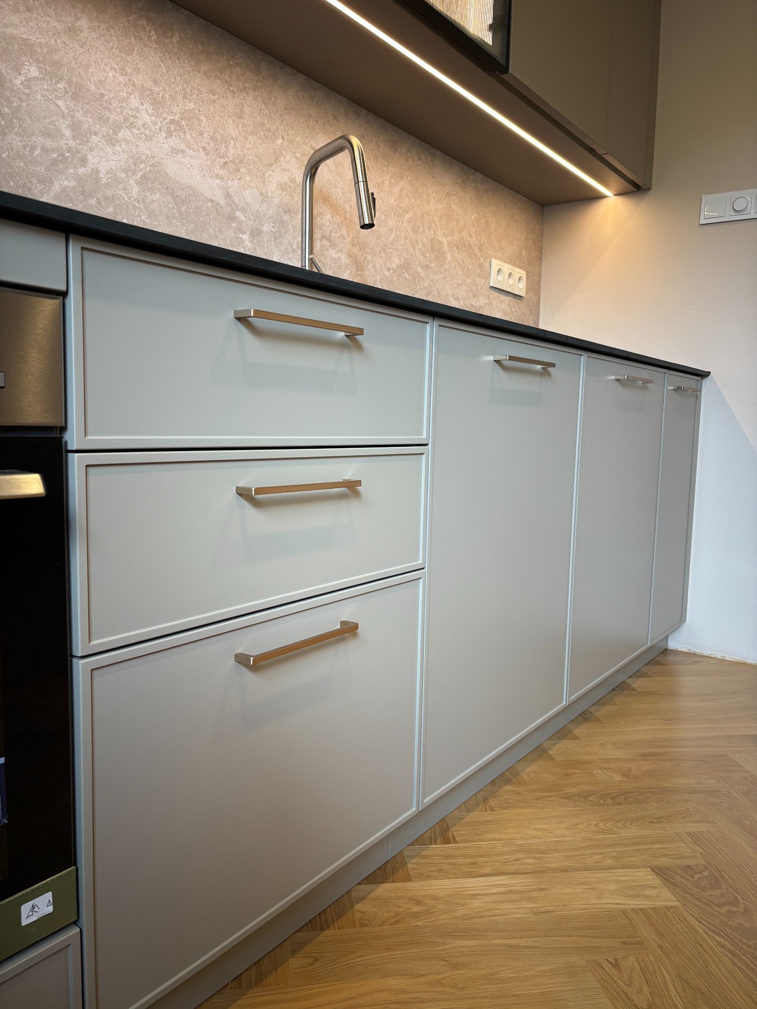 Szare szafki kuchenne z złotymi uchwytami, czarny blat i marmurowa ściana. Widoczny fragment piekarnika i parkiet w jodełkę. Nowoczesna kuchnia z oświetleniem LED.