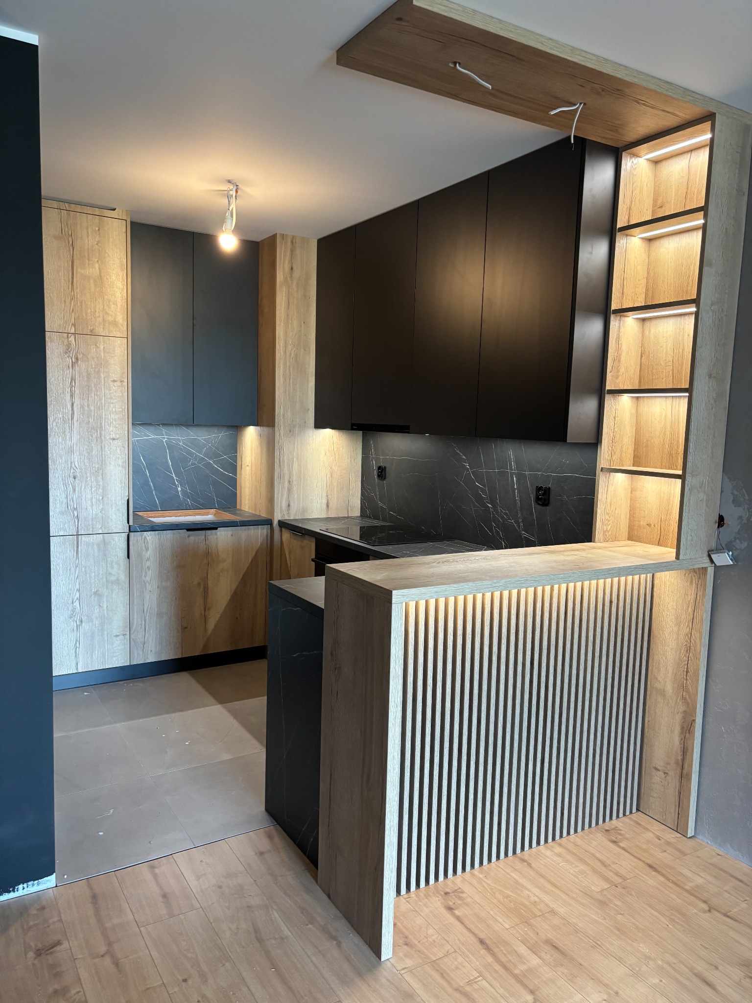 Nowoczesna kuchnia z drewnianymi i czarnymi frontami, marmurowym blatem i oświetleniem LED, w trakcie montażu. Widoczne niedokończone wykończenia ścian.