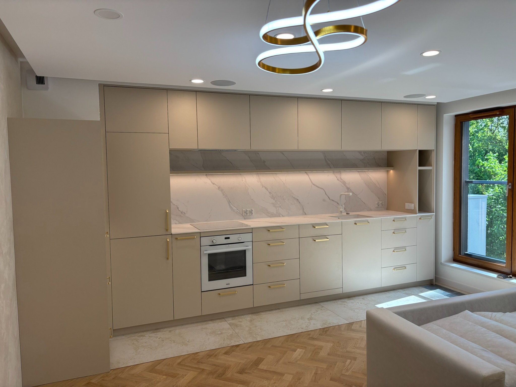 Jasna, nowoczesna kuchnia z beżowymi frontami, złotymi uchwytami i marmurowym blatem, oświetlona ledowym żyrandolem i światłem dziennym z okna.