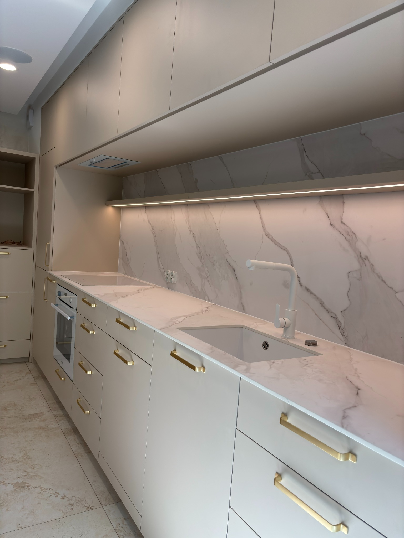 Jasna kuchnia z marmurowym blatem i złotymi uchwytami. Nowoczesny design z oświetleniem LED i minimalistyczną armaturą. Ujęcie z perspektywy podłogi.