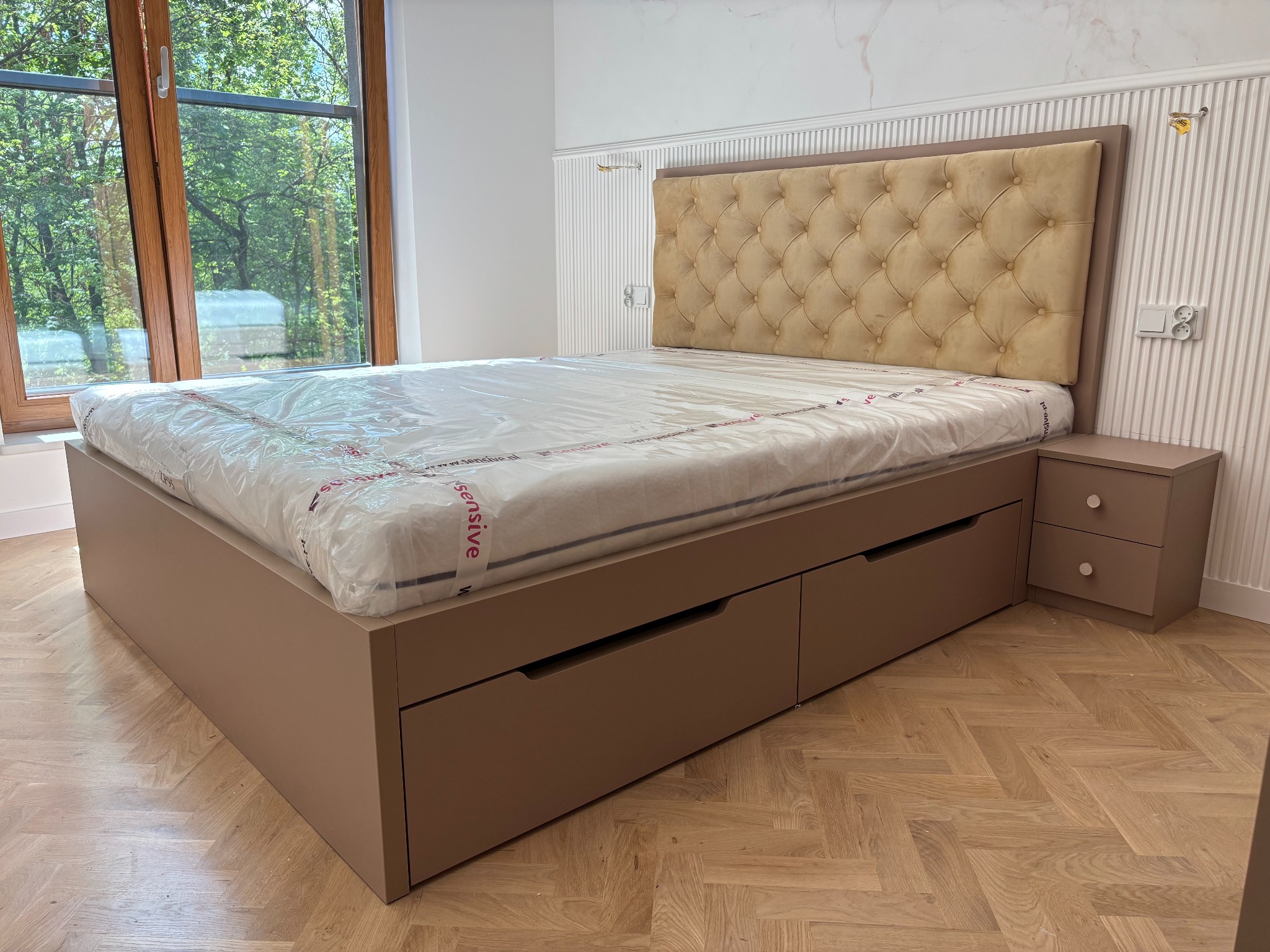 Beżowe łóżko na wymiar z pikowanym zagłówkiem i szufladami, z szafką nocną w tym samym kolorze, na tle jasnego wnętrza z oknem i parkietem.