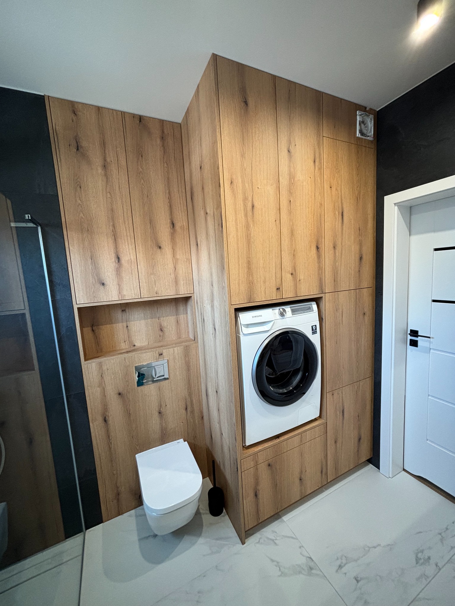 Drewniana zabudowa w łazience z pralką Samsung, toaletą i otwartą półką. Ciemne ściany, biała podłoga z marmurkowym wzorem, białe drzwi.