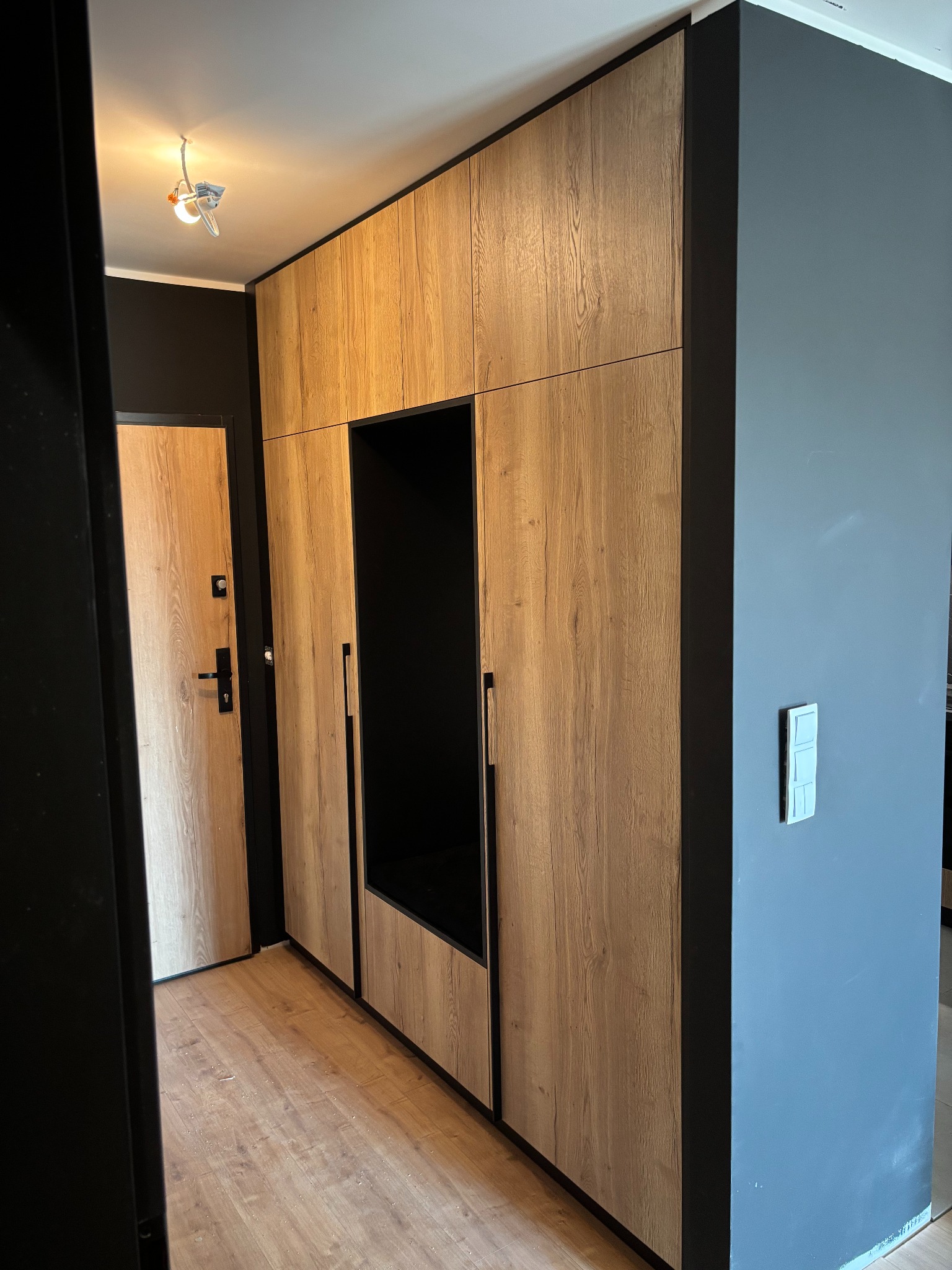 Zabudowa wnękowa z drewnopodobnymi frontami i czarnymi detalami, wkomponowana w przedpokój z drzwiami o podobnym wykończeniu i szarą ścianą.
