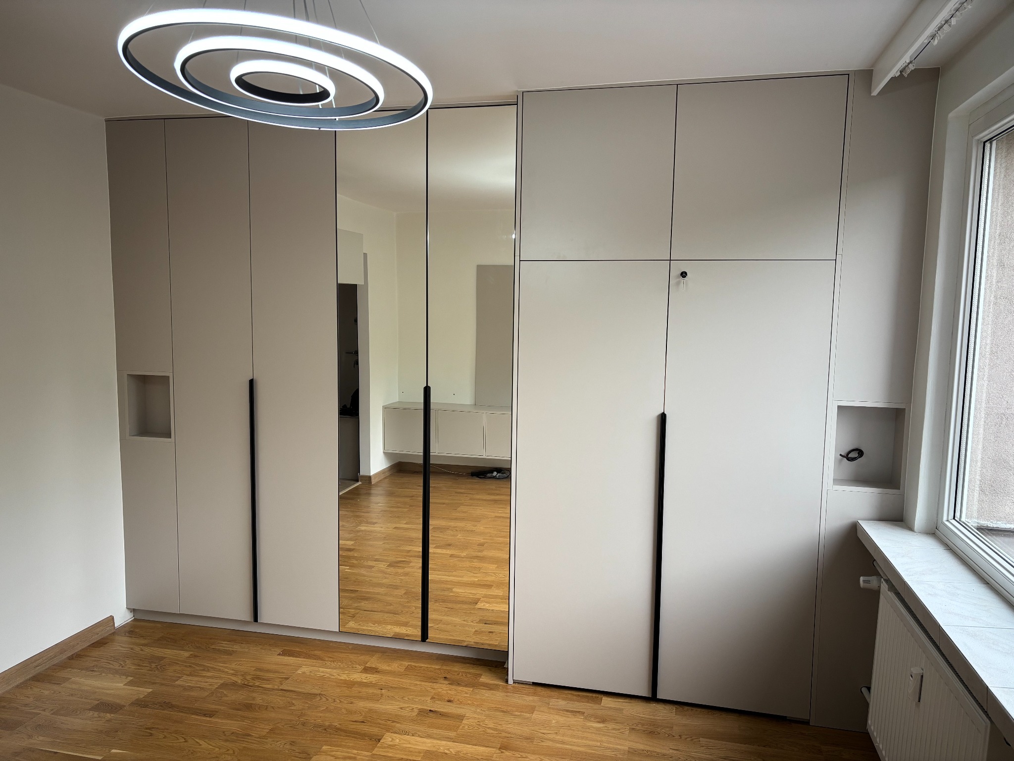 Jasna szafa wnękowa z lustrzanymi drzwiami, czarne, długie uchwyty, drewniana podłoga, widoczny fragment okna i grzejnika, nowoczesna lampa sufitowa.