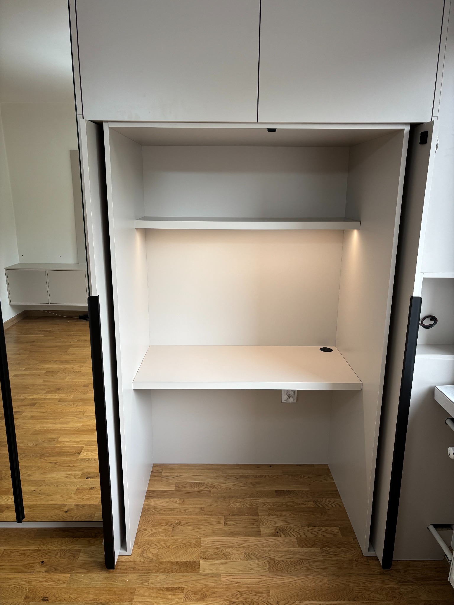 Biała szafa wnękowa z wysuwanym biurkiem, oświetleniem LED i gniazdkiem elektrycznym. Widoczne lustrzane drzwi przesuwne i drewniana podłoga.