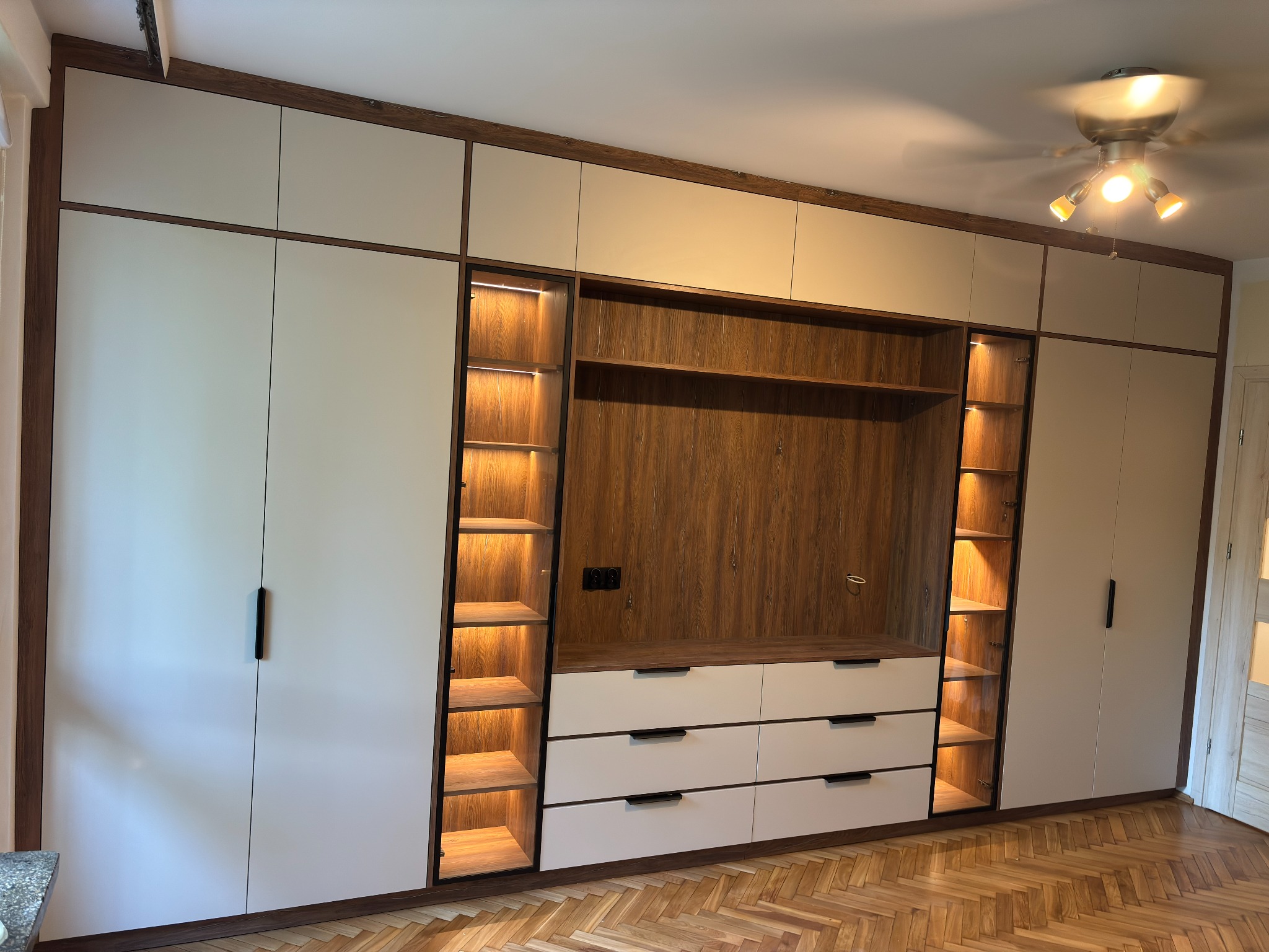 Biało-drewniana szafa wnękowa z oświetleniem LED półek, widok z przodu na parkiet w jodełkę. Minimalistyczne uchwyty, ciepłe światło lampy sufitowej.