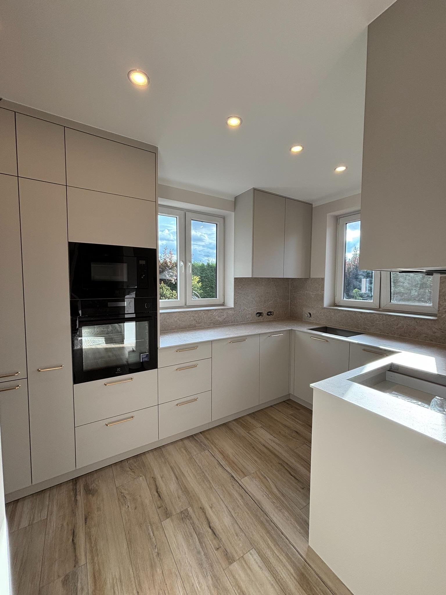 Jasna kuchnia z drewnianą podłogą, zabudową pod sufit i sprzętem AGD w zabudowie. Okno z widokiem na zieleń. Minimalistyczny design z złotymi uchwytami.