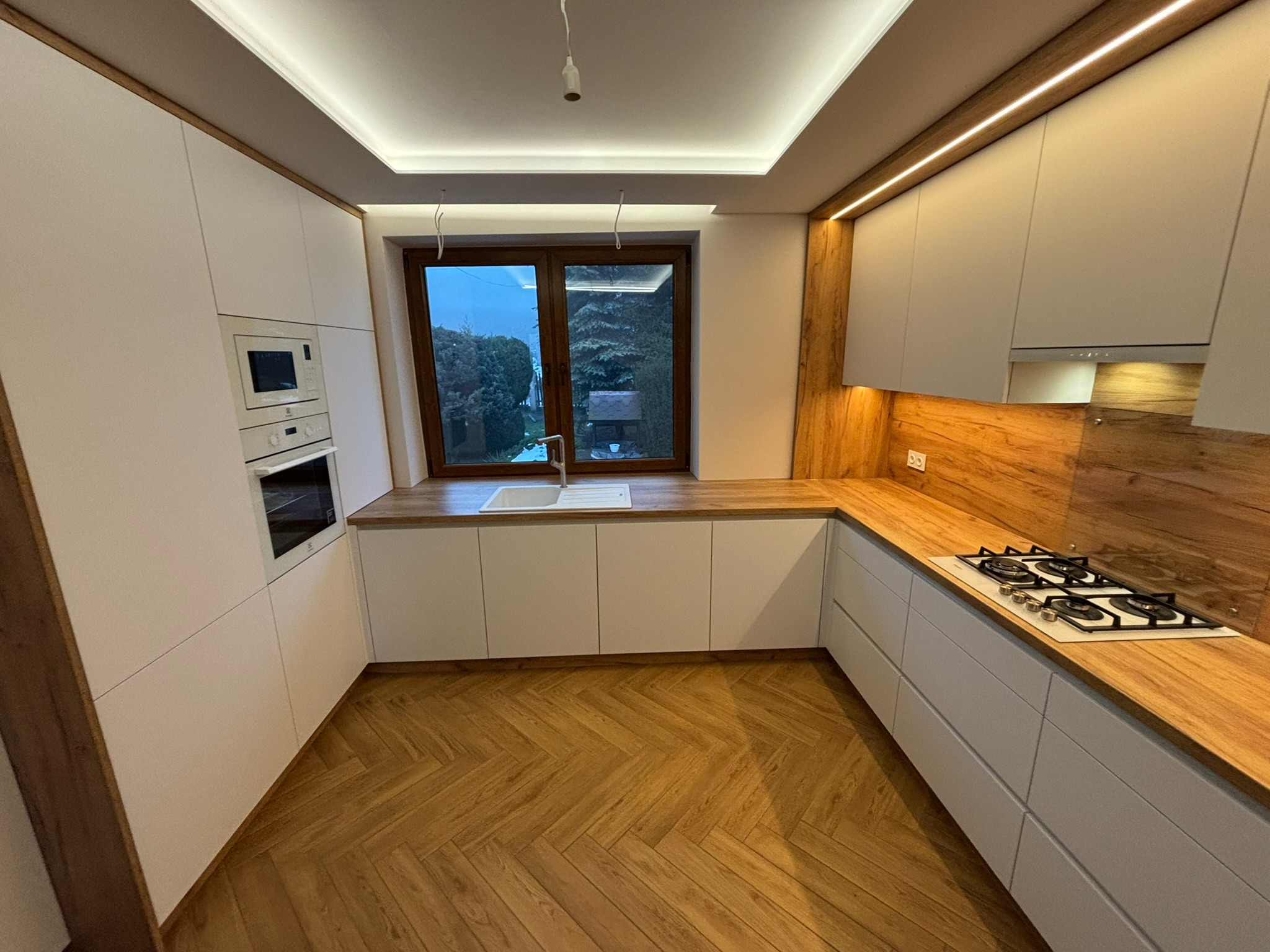 Nowoczesna kuchnia z białymi frontami, drewnianym blatem i podłogą w jodełkę. Widoczne oświetlenie LED podszafkowe i okno z widokiem na zieleń.