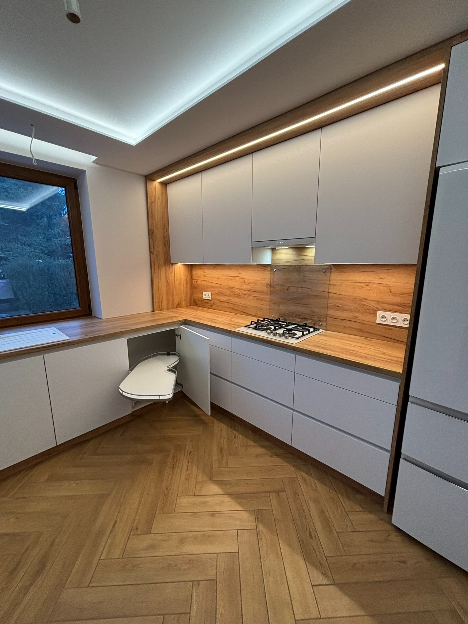 Nowoczesna kuchnia w Krakowie z szarymi frontami i drewnianym blatem. Widoczny system narożny z półkami obrotowymi. Oświetlenie LED podkreśla detale.