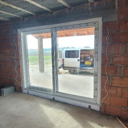 Adam Kucal - Montaż przesuwnych okien w surowym stanie, widoczny mur z cegieł, na zewnątrz biały bus z narzędziami.