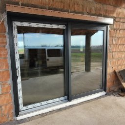 Adam Kucal - Montaż okna przesuwnego z roletą zewnętrzną w surowym stanie budynku z cegły, widoczne narzędzia i osoba w tle.