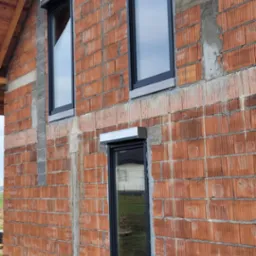 Fragment ściany domu w trakcie budowy z zamontowanymi oknami PCV w czarnych ramach i roletami zewnętrznymi z aluminiowymi kasetami w kolorze srebrnym, widoczne niedokończone spoiny między cegłami.