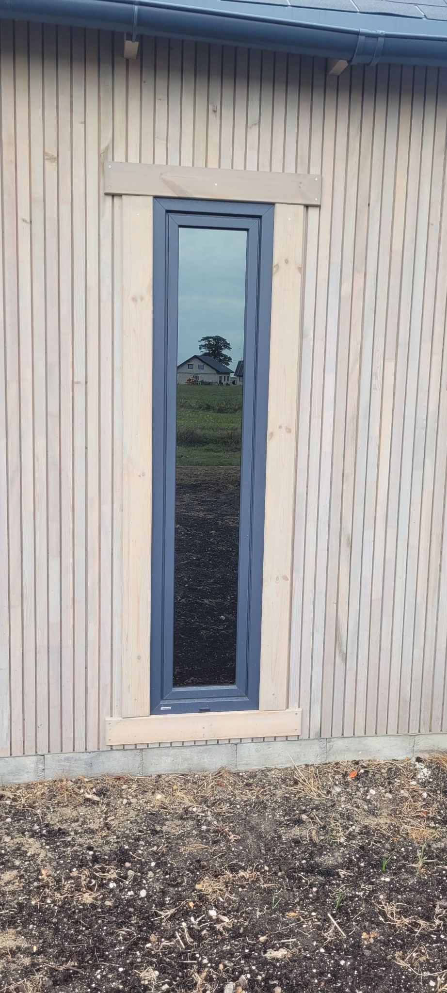 Wąskie okno z szarą ramą zamontowane w elewacji z jasnego drewna, w szybie odbija się dom i drzewo na tle pochmurnego nieba.