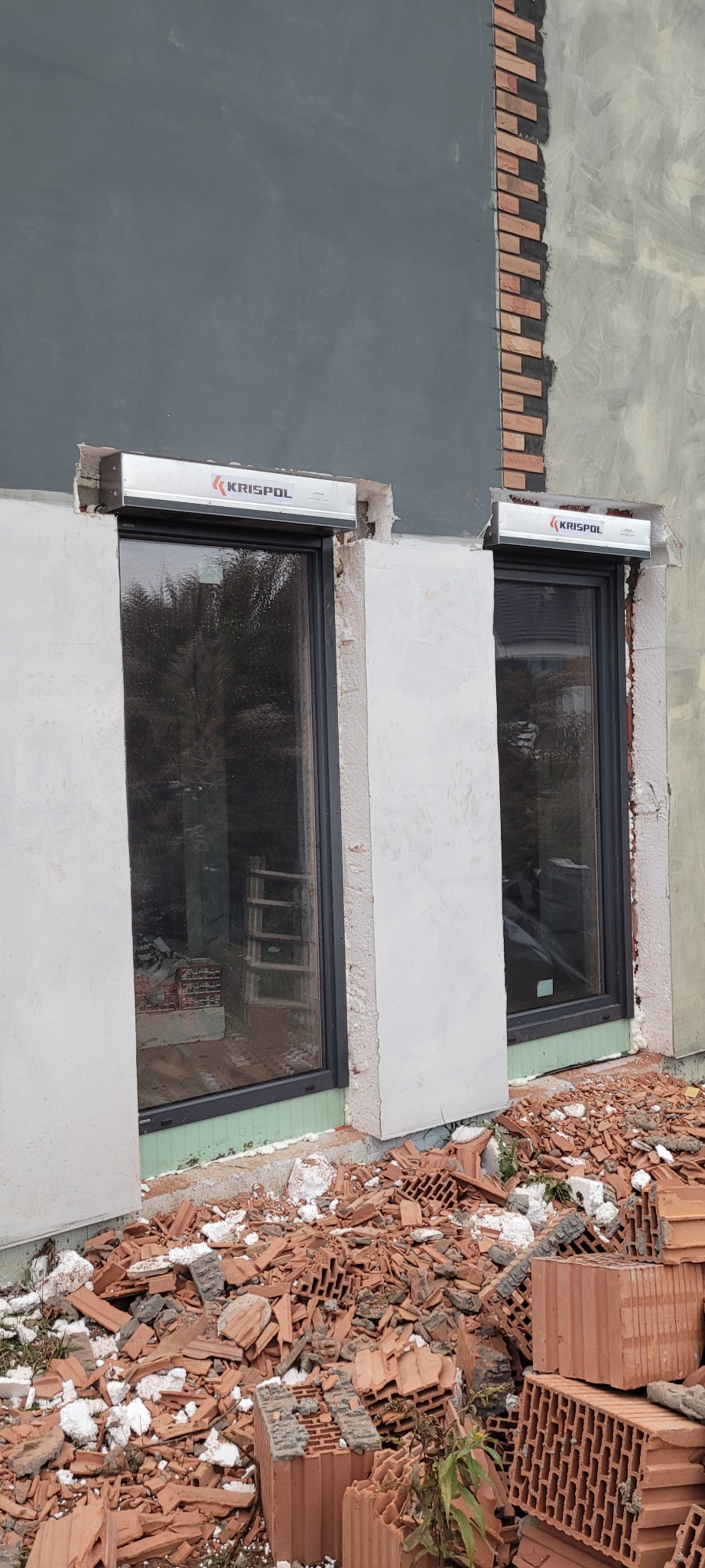 Dwa okna z zamontowanymi roletami zewnętrznymi marki Krispol w trakcie montażu, widoczne niedokończone elewacje i gruz ceglany.