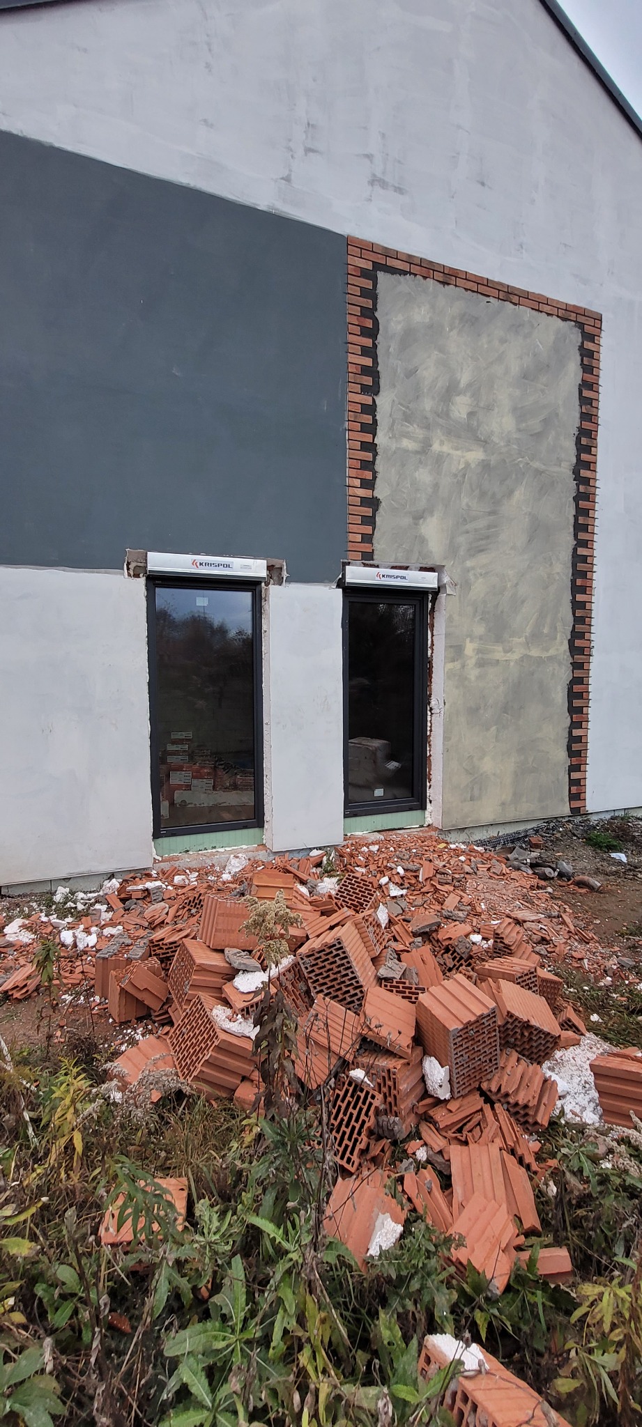 Montaż stolarki okiennej w trakcie prac wykończeniowych elewacji budynku, widoczne czarne ramy okienne z roletami zewnętrznymi i sterta gruzu ceglanego przed oknami.