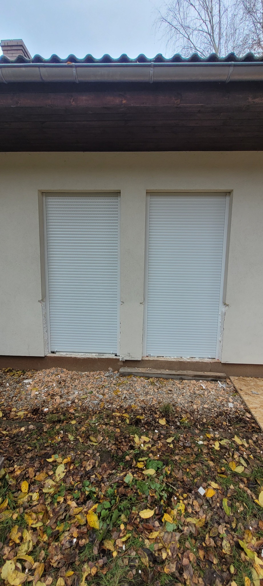 Dwa okna z opuszczonymi, białymi roletami zewnętrznymi na elewacji budynku, widoczne ślady prac budowlanych na dole.