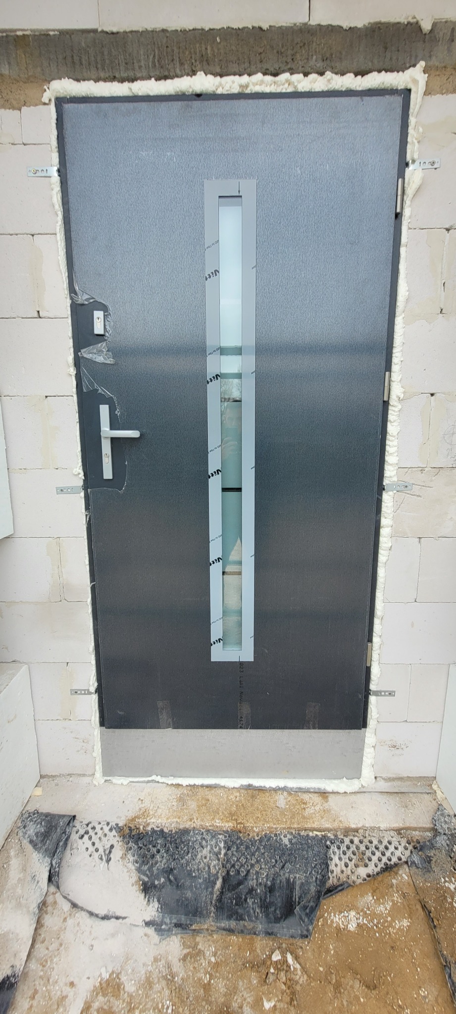 Montaż szarych drzwi zewnętrznych z długim, pionowym przeszkleniem, widoczne pianowanie i elementy montażowe, surowy stan wykończenia wnętrza.
