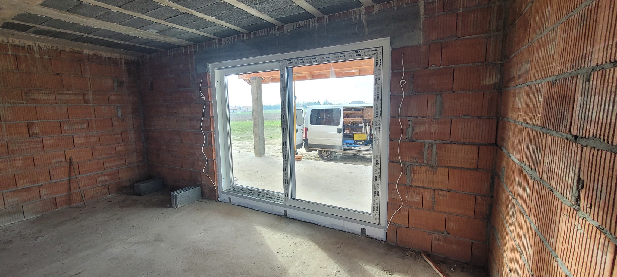 Montaż przesuwnych okien w surowym stanie, widoczny mur z cegieł, na zewnątrz biały bus z narzędziami.