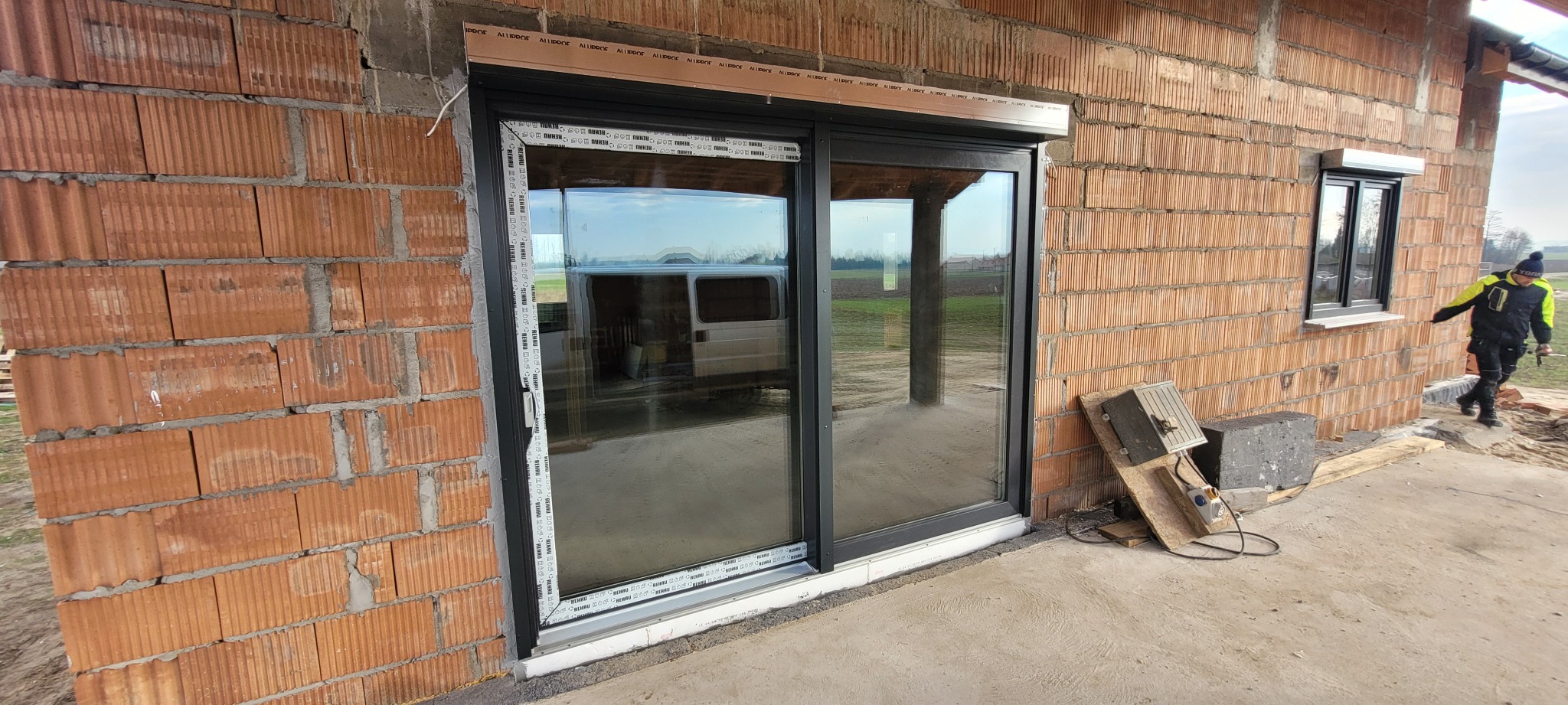 Montaż okna przesuwnego z roletą zewnętrzną w surowym stanie budynku z cegły, widoczne narzędzia i osoba w tle.