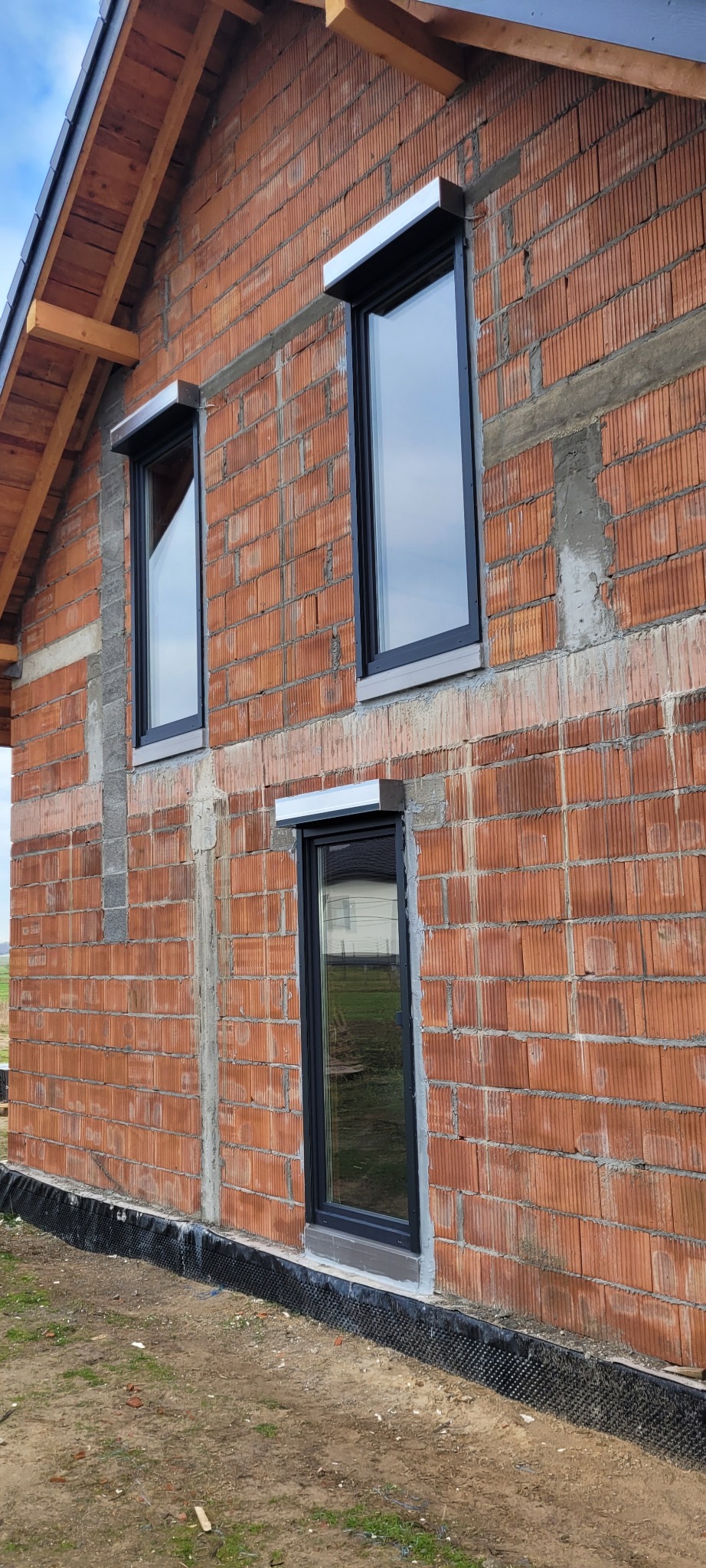 Fragment ściany domu w trakcie budowy z zamontowanymi oknami PCV w czarnych ramach i roletami zewnętrznymi z aluminiowymi kasetami w kolorze srebrnym, widoczne niedokończone spoiny między cegłami.