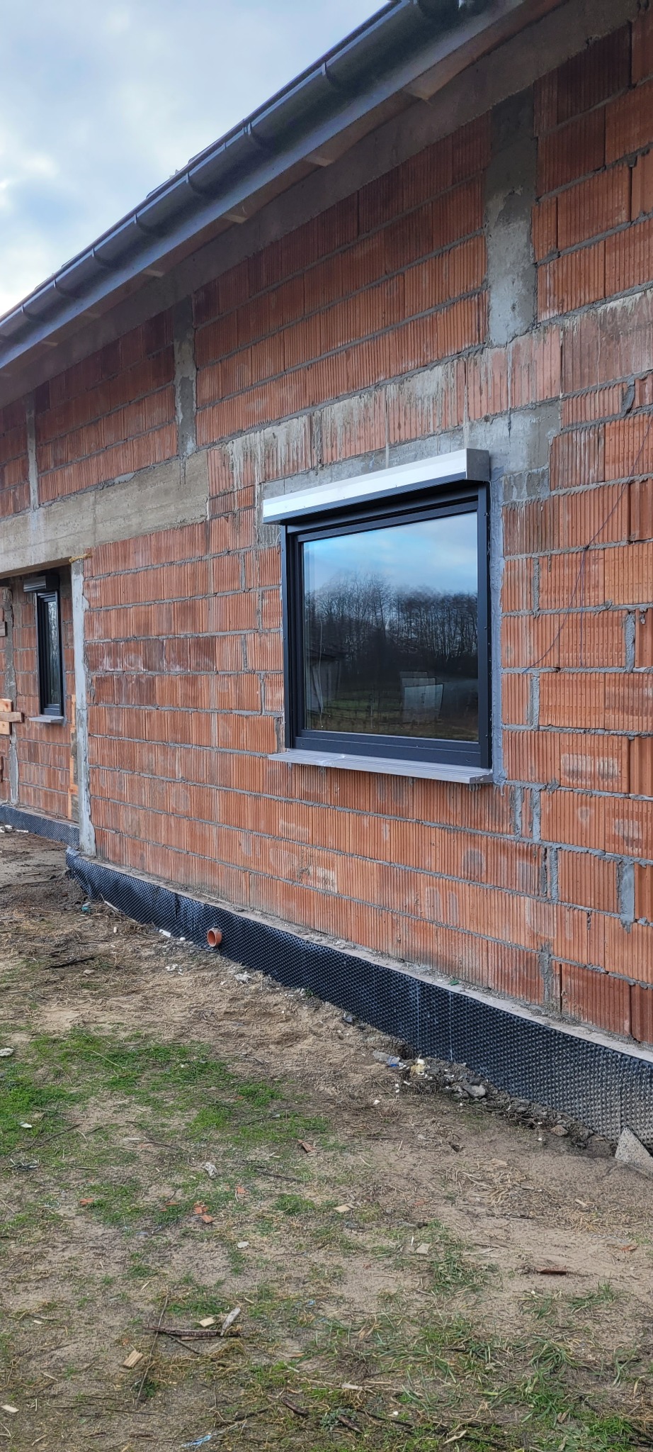 Surowy mur z czerwonej cegły z zamontowanym oknem z czarną ramą i szarą roletą zewnętrzną, w tle odbicie drzew i nieba.