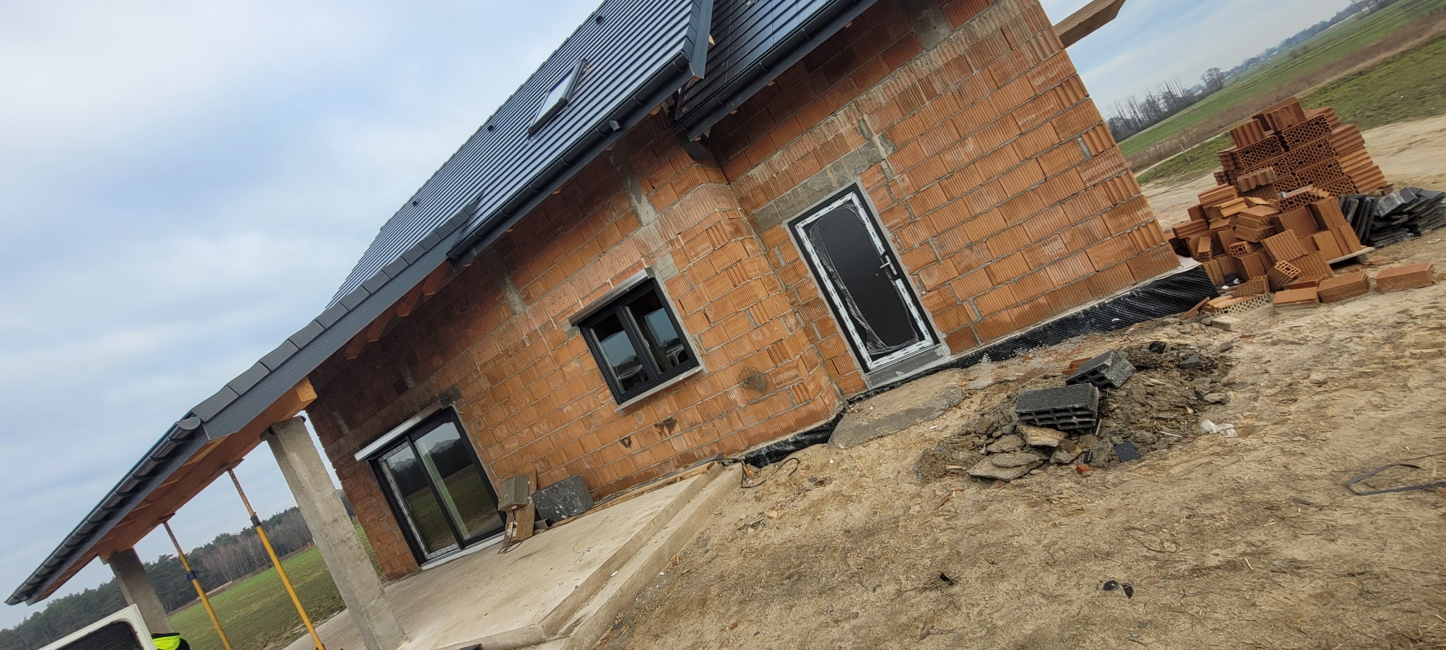 Dom jednorodzinny w trakcie budowy, widoczne czarne ramy okienne i drzwi wejściowe zabezpieczone folią, ściany z czerwonej cegły, szare dachówki, teren wokół domu w stanie surowym z gruzem i stertą...