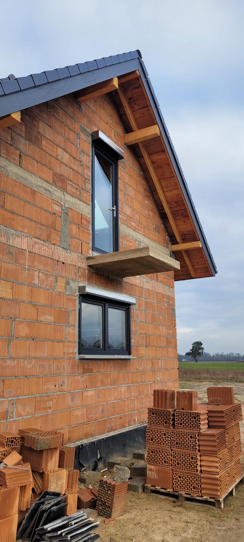 Budynek w trakcie budowy z zamontowanymi oknami z roletami zewnętrznymi, widoczne cegły i drewniana konstrukcja dachu.