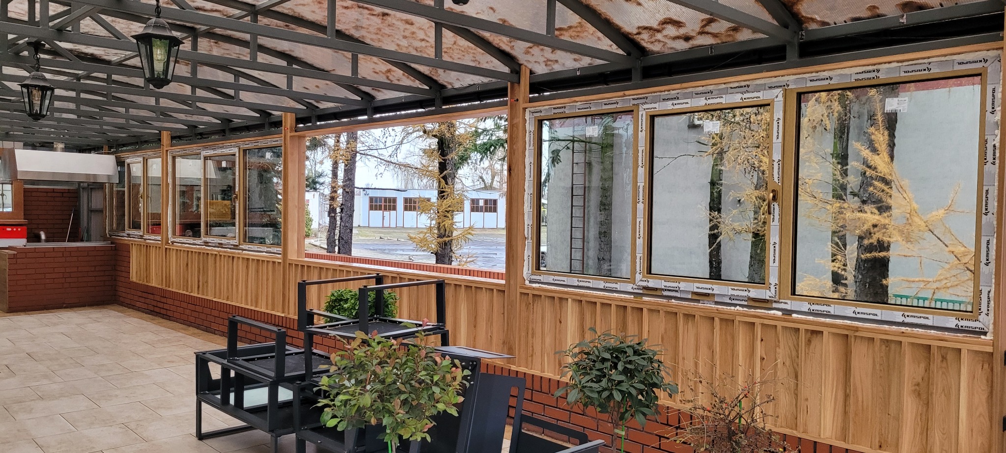 Zadaszony taras z drewnianą boazerią i nowymi oknami z Izbicy Kujawskiej, w których odbijają się drzewa i budynek gospodarczy.