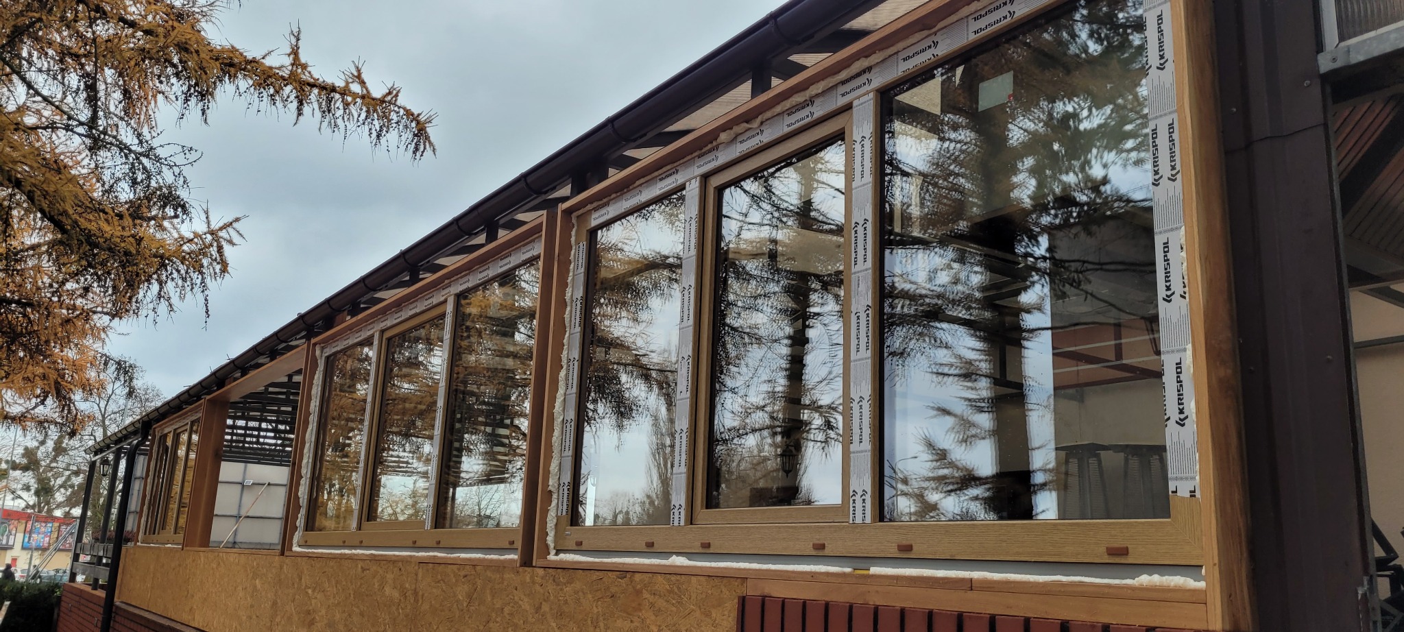 Zewnętrzny widok przeszklonej werandy z drewnianą ramą okienną, zabezpieczonej taśmą montażową z logo producenta, odbijającej drzewa na tle pochmurnego nieba.
