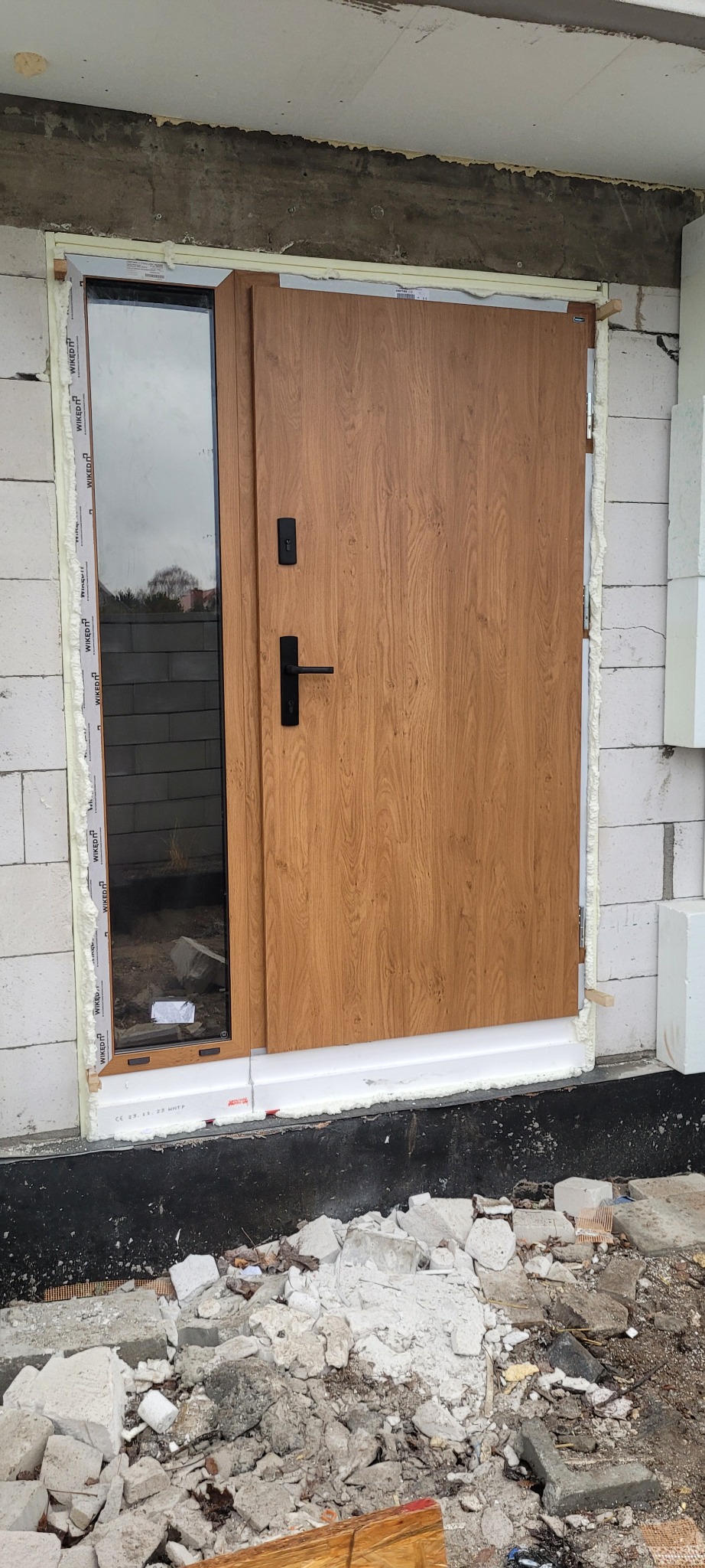 Montaż drzwi zewnętrznych z bocznym oknem w surowym stanie, widoczne bloki betonowe, pianka montażowa i gruz budowlany.