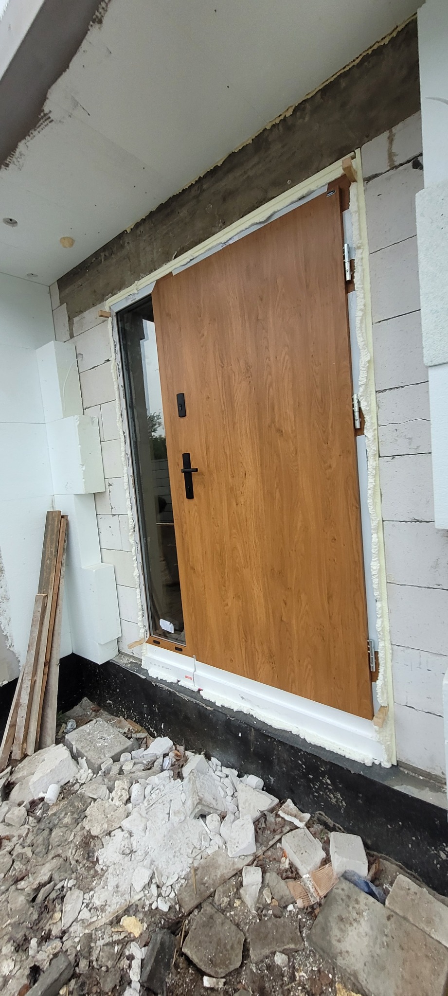 Montaż brązowych drzwi zewnętrznych z czarną klamką w surowym stanie, widoczne białe bloczki i pianka montażowa.