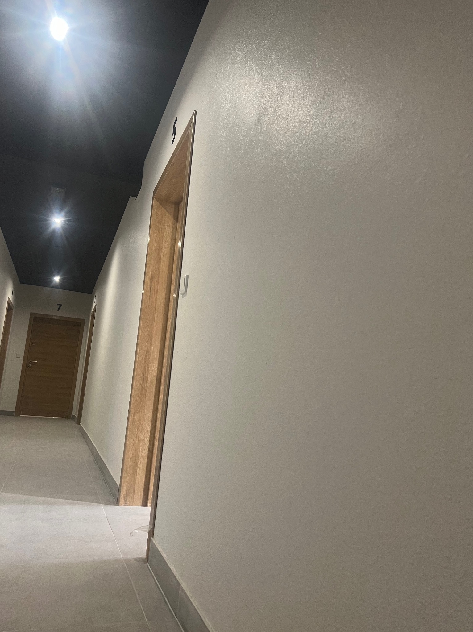 Korytarz w budynku z numerami pokoi oświetlony punktowymi lampami sufitowymi, ściany pomalowane na jasny kolor, widoczna faktura gładzi.